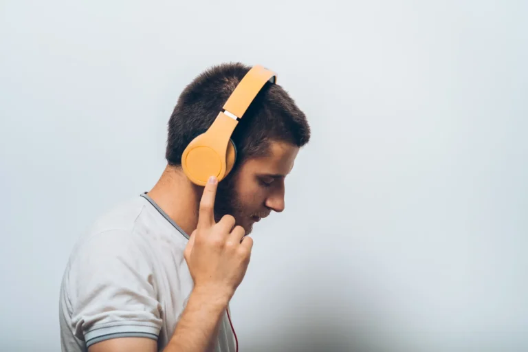 Young man wearing bright orange over-ear headphones, listening intently with eyes closed and finger touching the side of the headset.