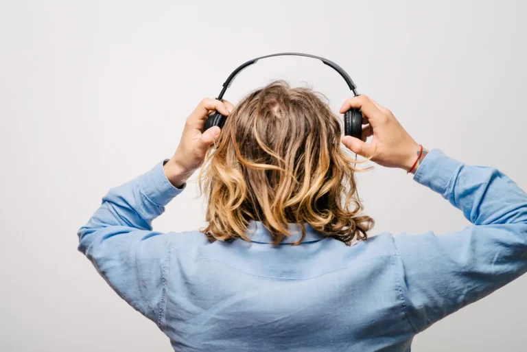 Person with long wavy hair and a blue shirt seen from behind, placing black headphones over their ears.